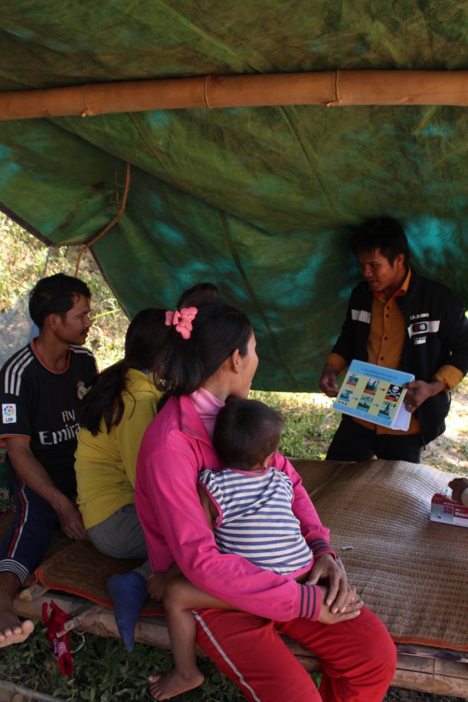 A family gathers in the shade under a tarpaulin to learn about the causes of malaria
