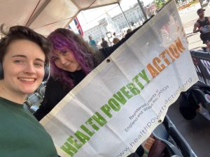 Two people smile at the camera, one holds a banner with 'health poverty action' written on the front.