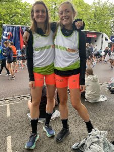 Two women with their arms around each other smile at the camera. They are wearing running vests with 'health poverty action' written on the front. 